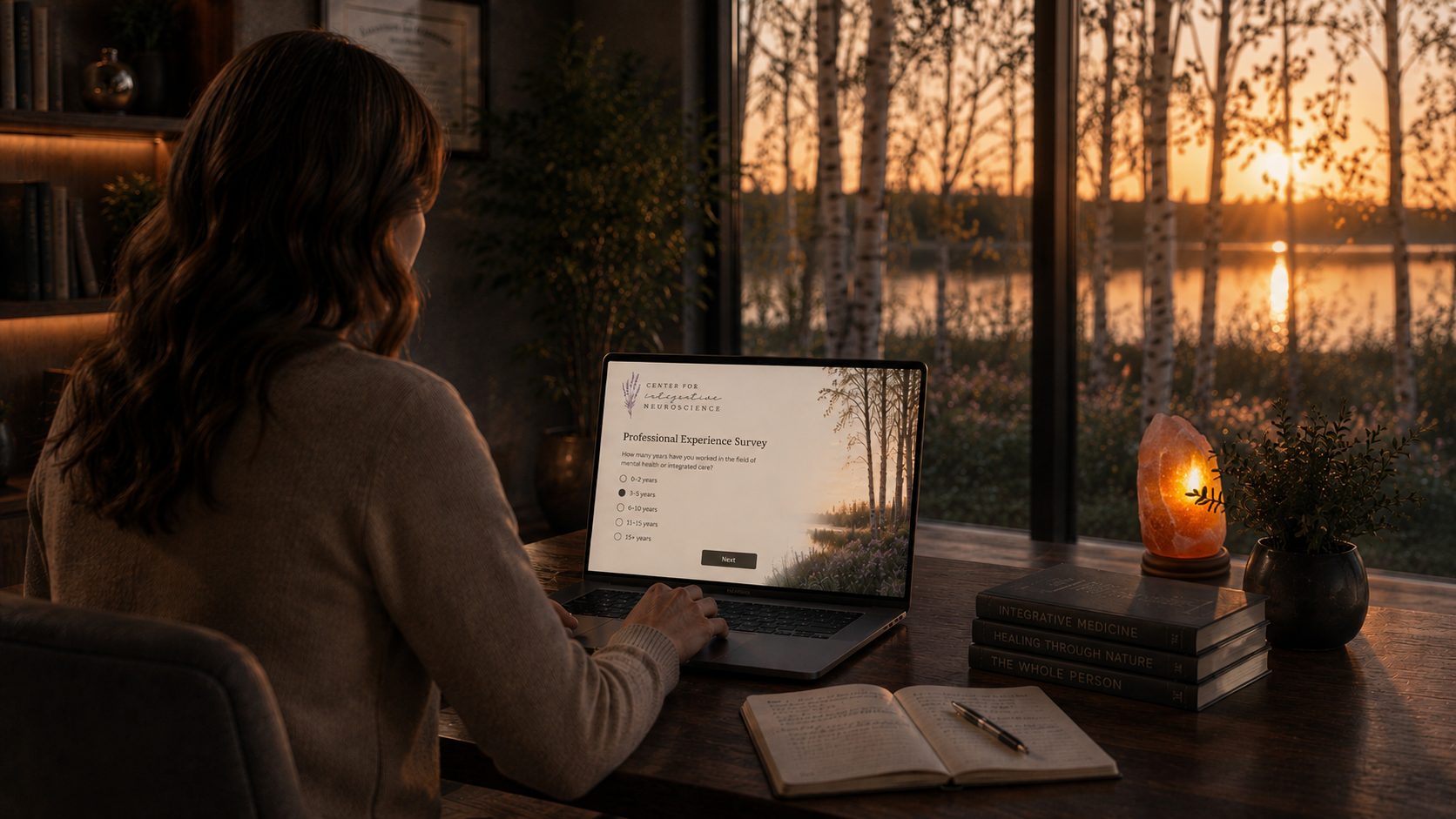 A clinician at a desk at sunset answering a Center for Integrative Neuroscience professional experience survey, a lake and birch trees beyond the window.